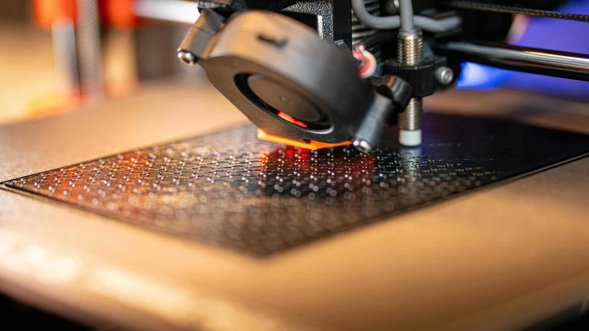 Macro view of a 3D printer nozzle actively extruding a textured layer of black polymer plastic filament, highlighting carbon-reinforced manufacturing materials.