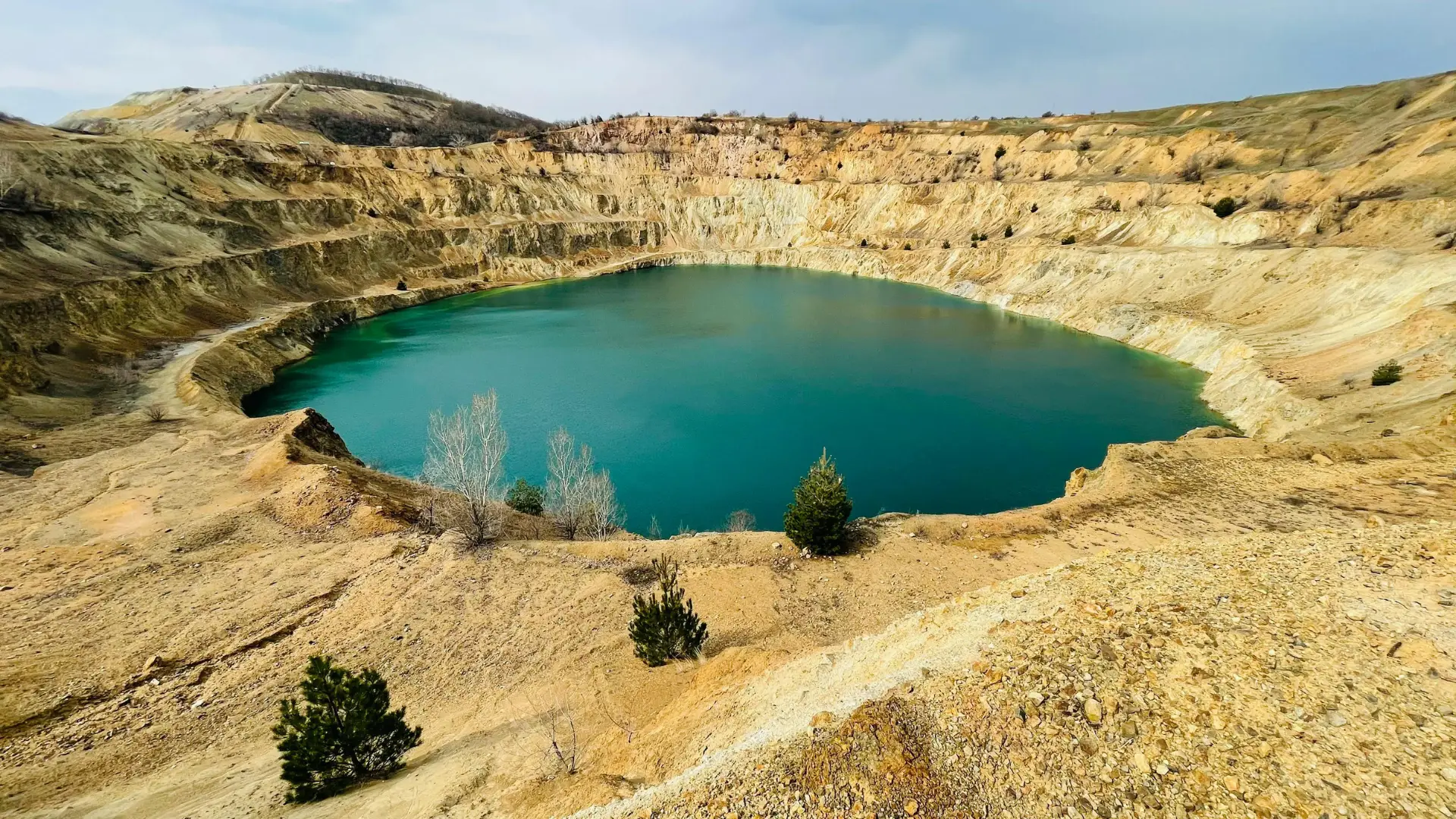 A massive, stepped open-pit mine crater that has filled with striking, deeply colored turquoise water, surrounded by an arid, rocky landscape.