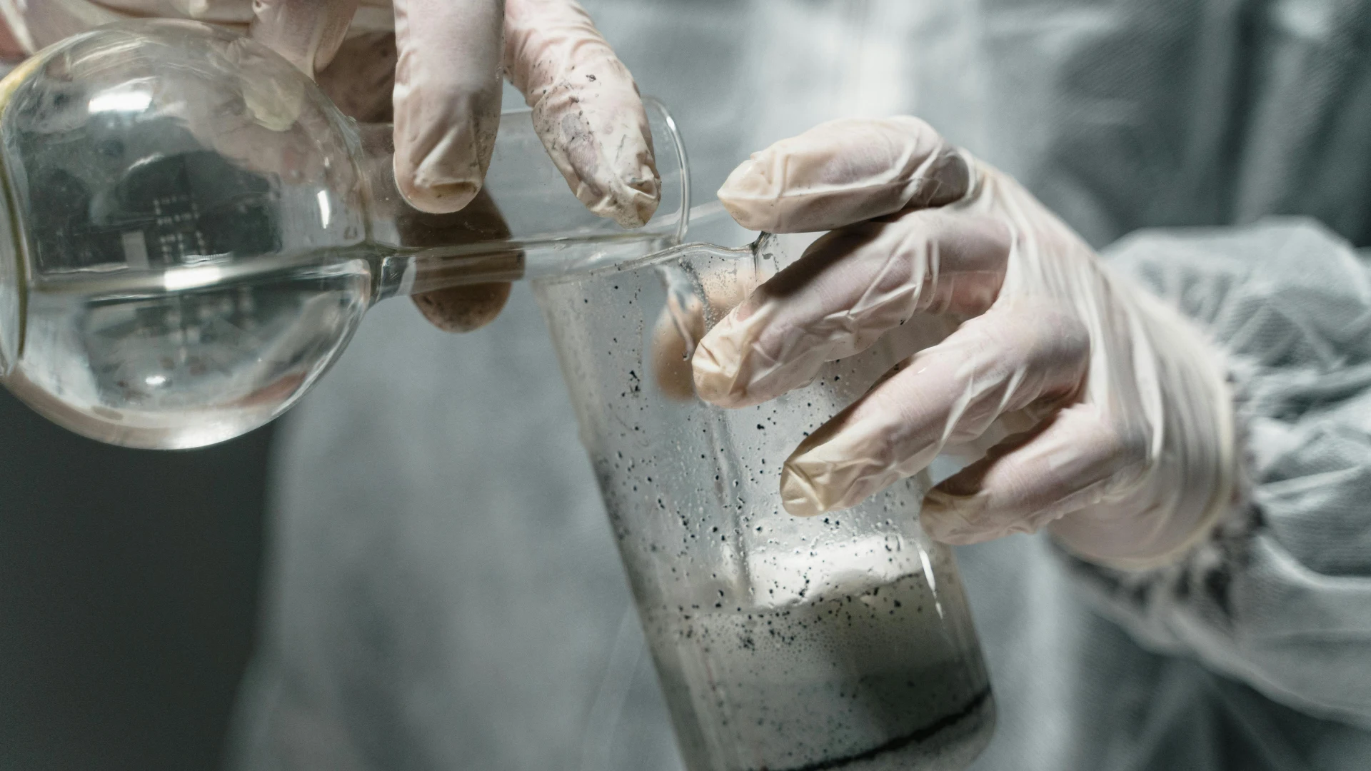 Laboratory technician in protective gloves pouring a clear chemical solution into a glass beaker containing black activated carbon powder for quality control and purification testing.