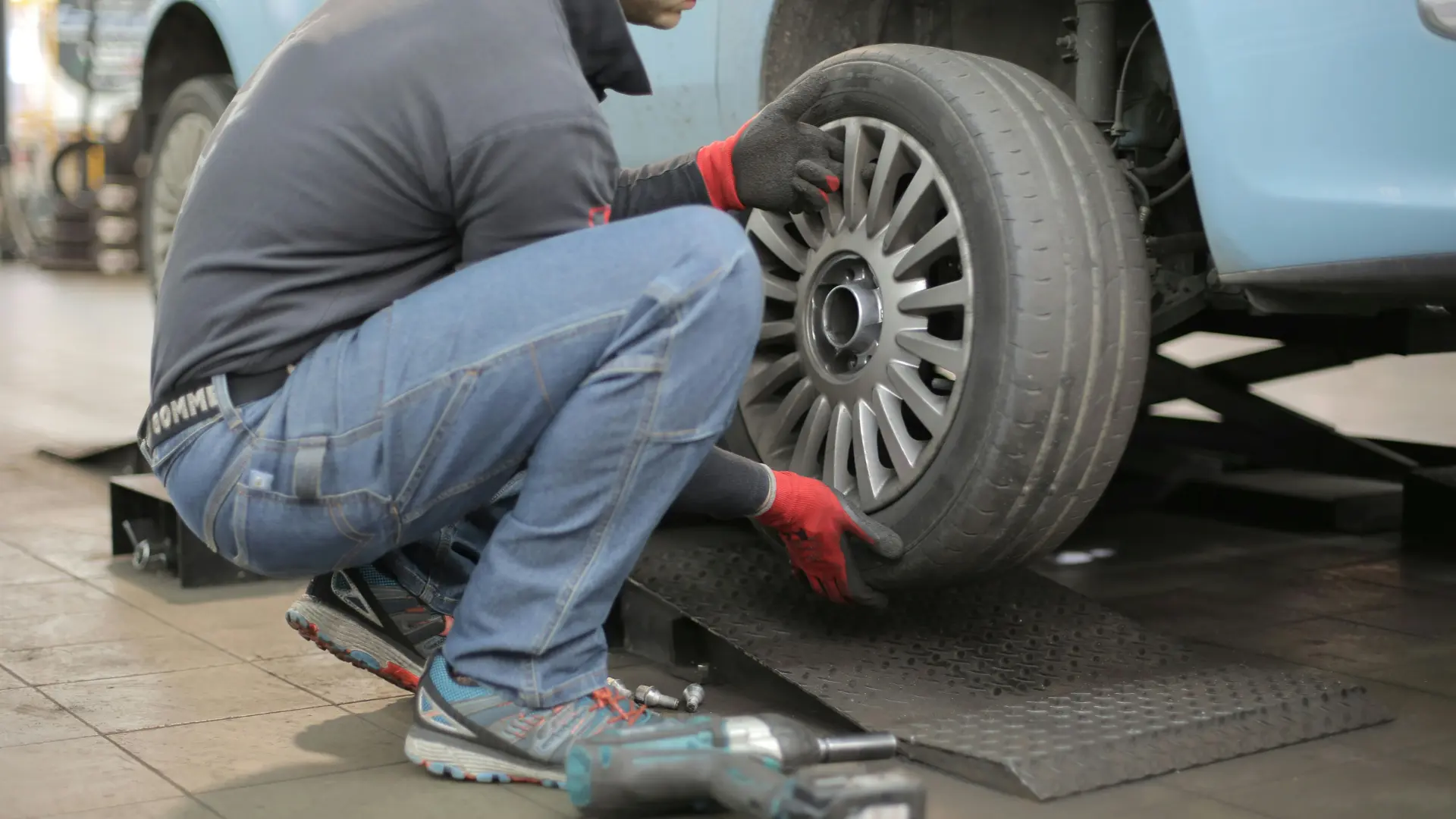 An automotive mechanic kneeling to remove or install a wheel and rubber tire on a blue vehicle elevated on a garage service lift.