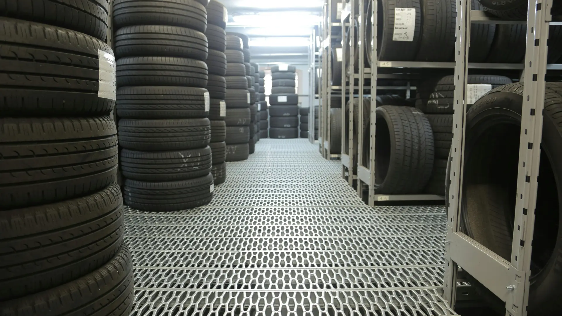 A brightly lit commercial auto shop warehouse featuring rows of new rubber vehicle tires neatly organized on metal grating and shelving racks.