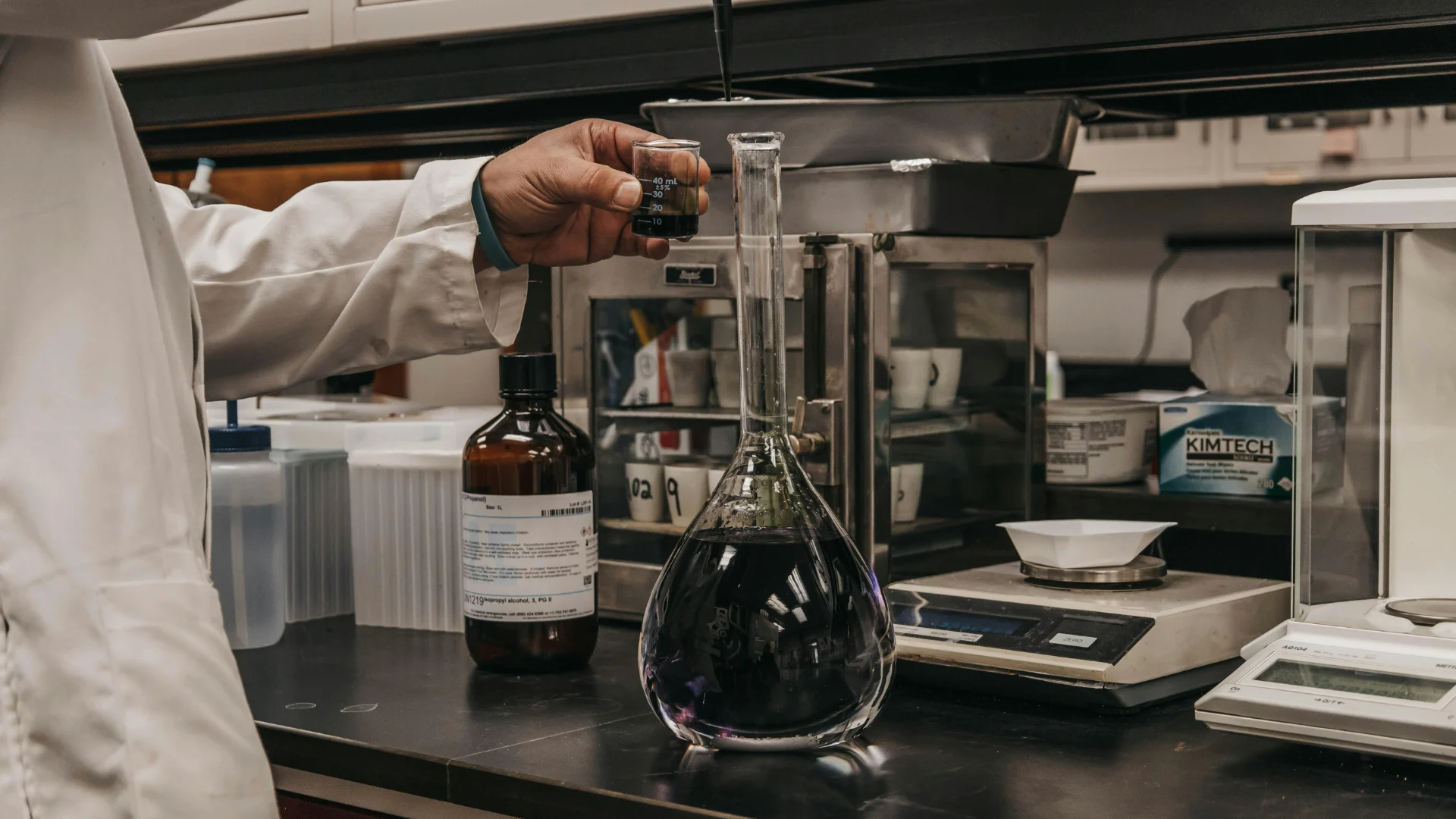 A laboratory technician in a white coat carefully measuring and pouring a dark, opaque chemical solution—such as a carbon black suspension—into a large glass flask for material analysis and quality control.