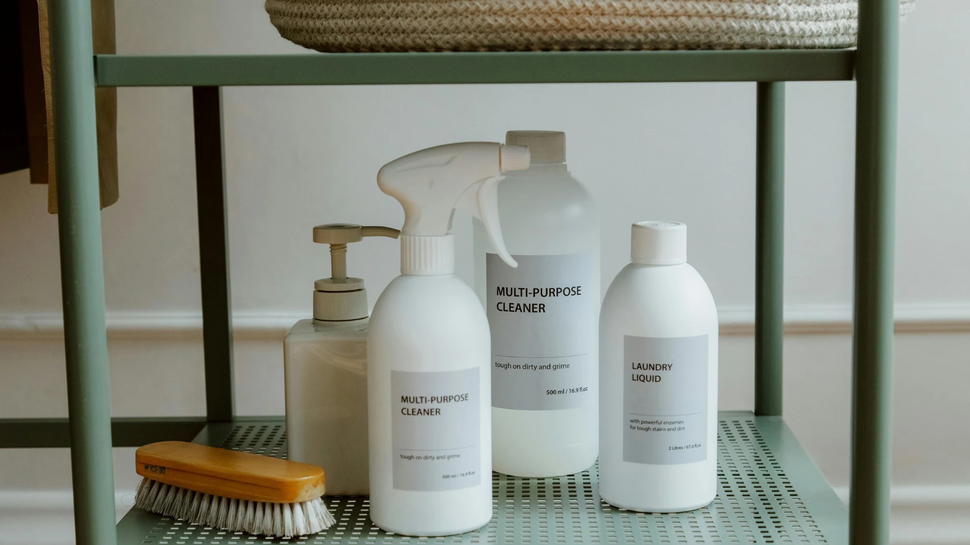 White plastic bottles of multi-purpose cleaner and laundry liquid on a green metal shelf, representing commercial chemical manufacturing, formulation, and packaging.