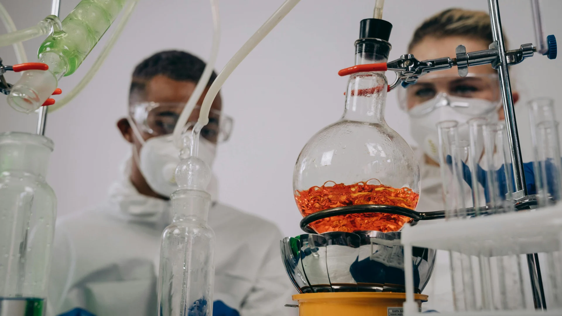 Two focused laboratory scientists wearing protective masks and safety goggles monitoring a complex glass distillation apparatus containing a boiling orange chemical liquid.