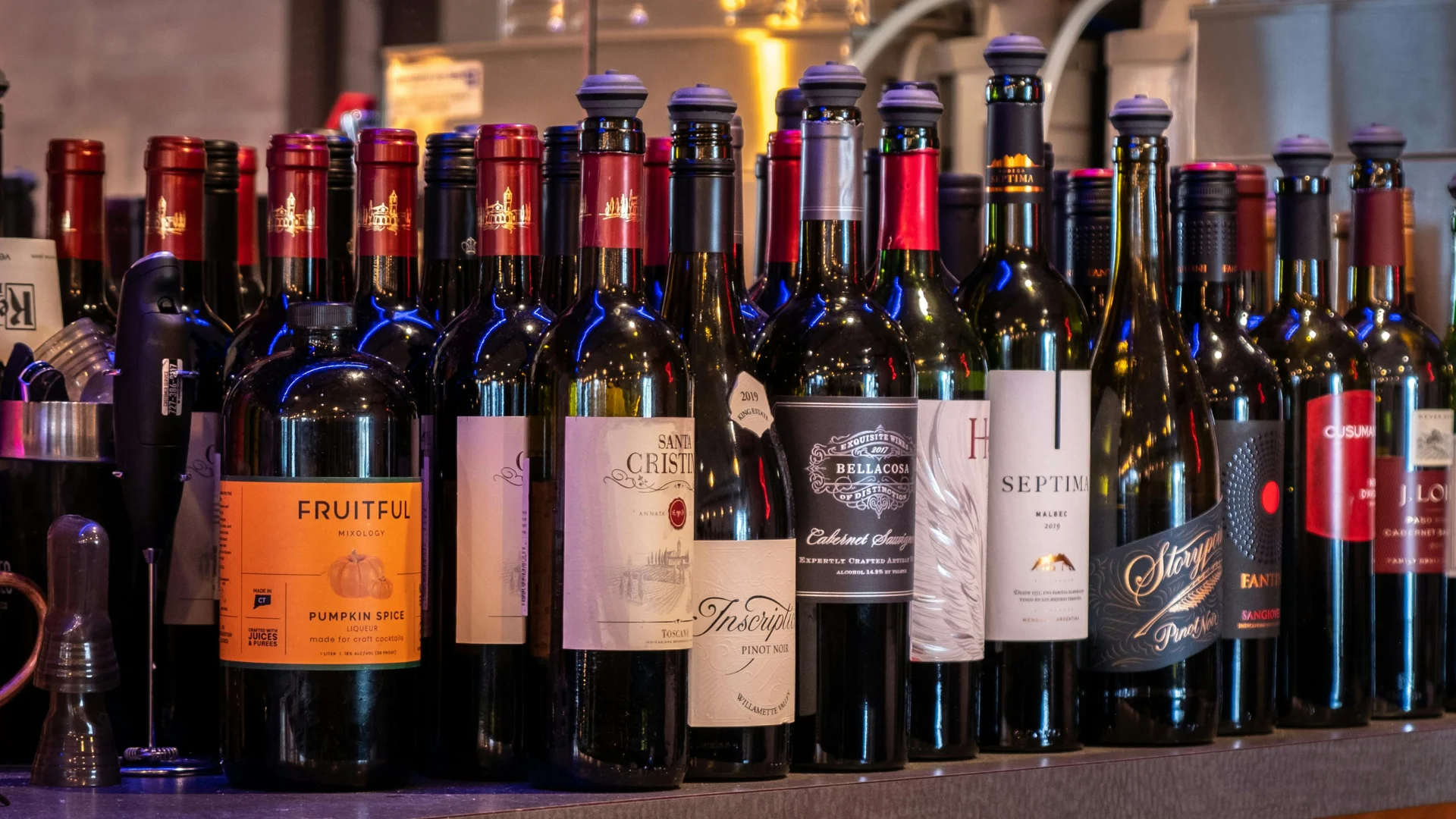 An assortment of commercial wine, liquor, and cocktail mixing syrup bottles arranged closely together on a dark bar counter setup.
