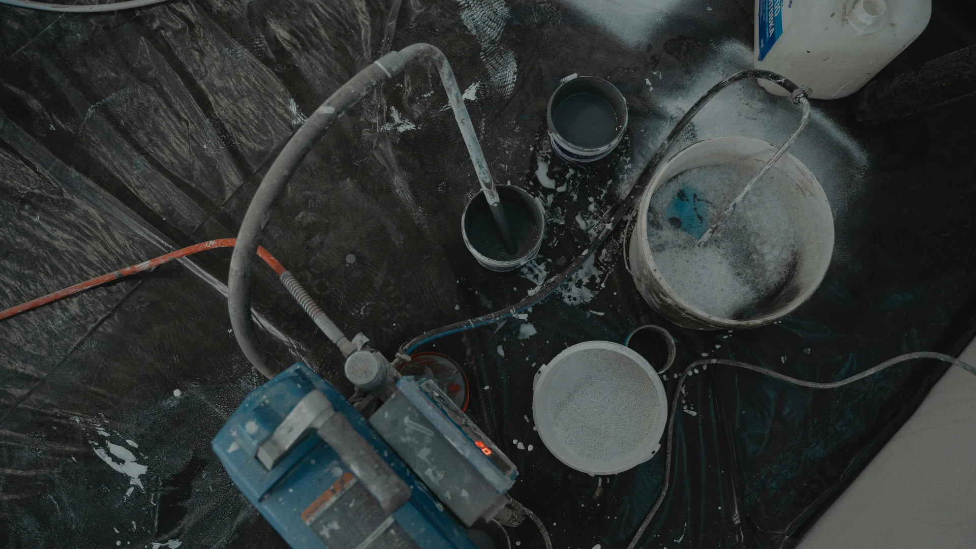 Top-down view of a messy industrial workspace featuring commercial paint spraying equipment, hoses, and mixing buckets used for heavy coatings and chemical pigment dispersion.