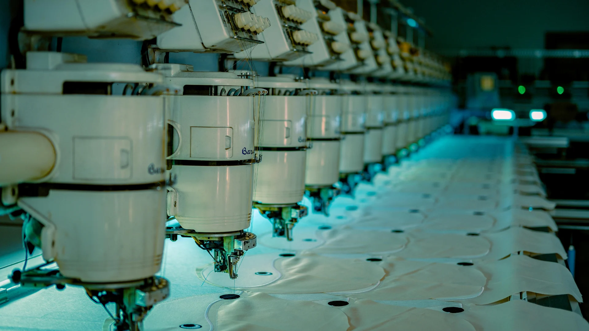 A long row of automated industrial embroidery and sewing machines operating in a large-scale commercial textile manufacturing facility.
