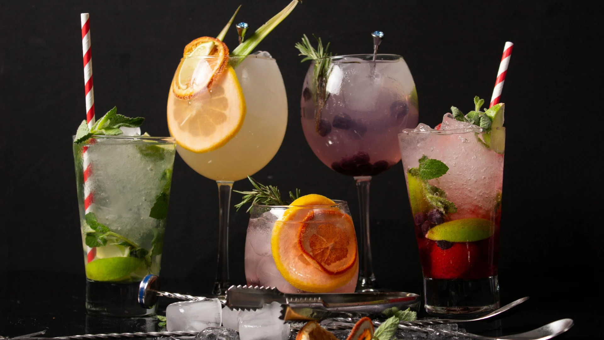 A diverse selection of brightly colored, refreshing craft cocktails and mocktails garnished with fresh citrus fruit, berries, and herbs on a dark background.