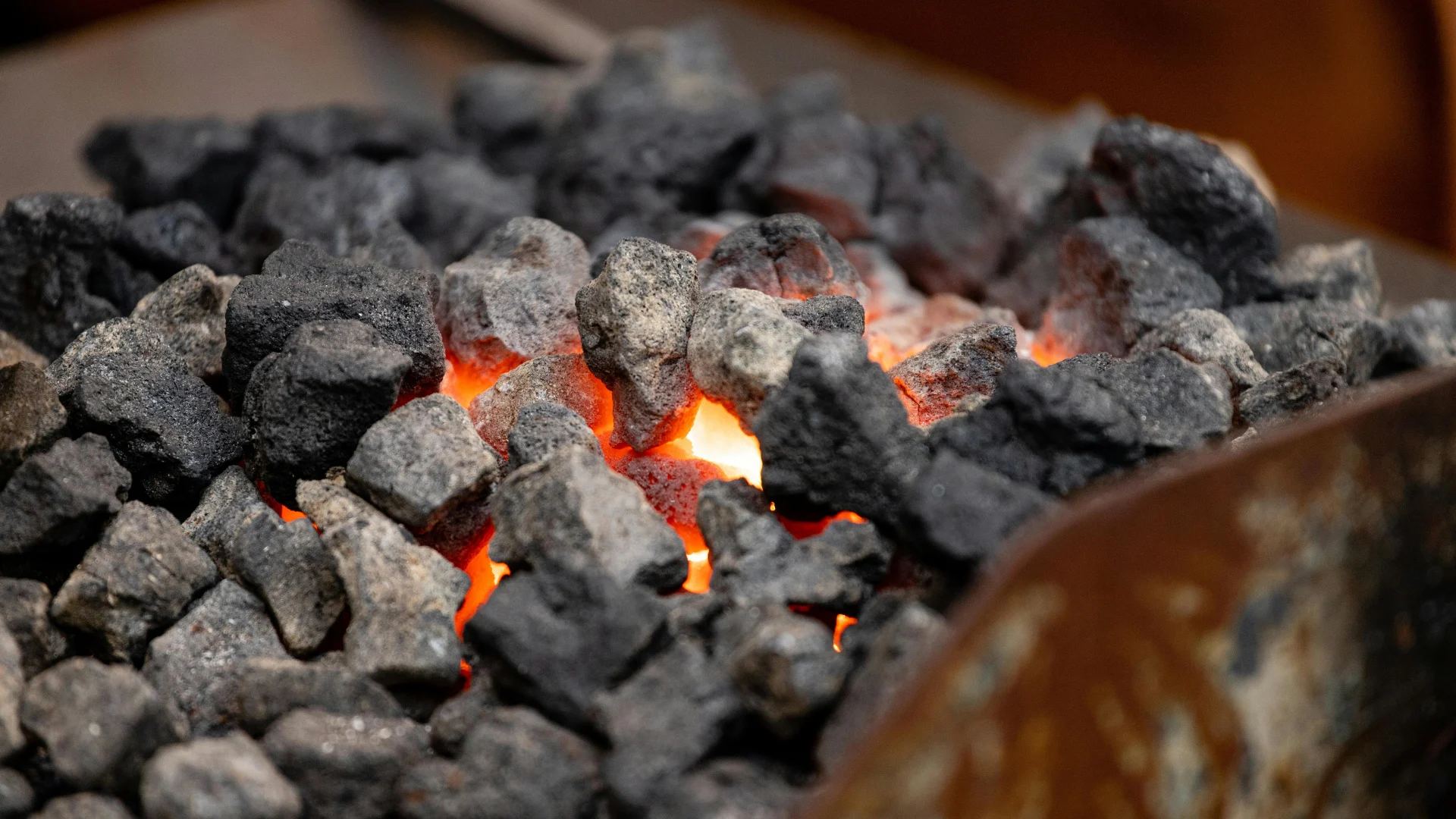 Close-up macro photography of rough black coal or natural lump charcoal pieces, with the center glowing an intense, high-heat red.