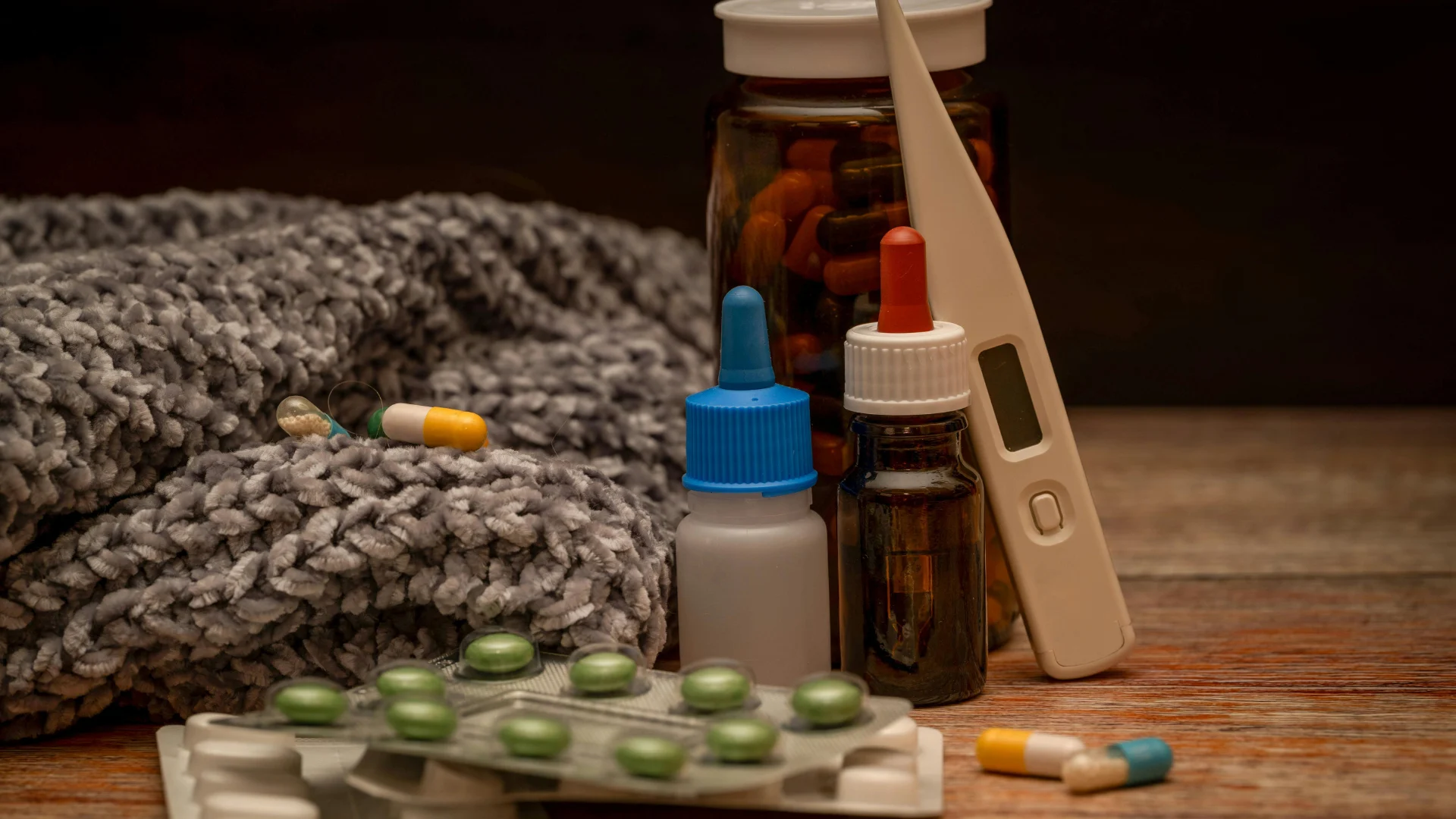A collection of home healthcare supplies including pill blister packs, liquid medicine dropper bottles, a digital thermometer, and a cozy gray blanket on a wooden table.