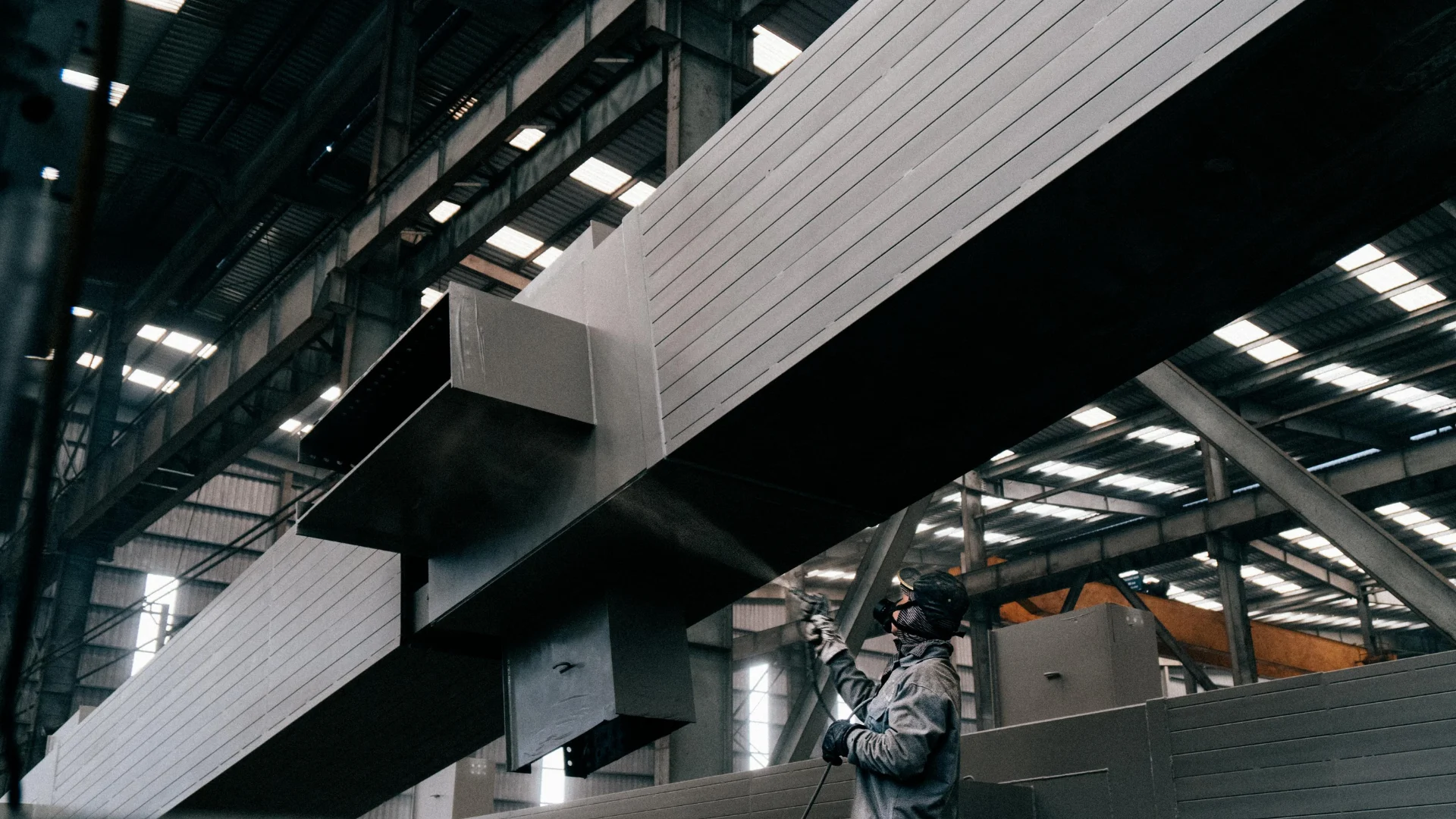 An industrial worker wearing safety respirator gear applying a heavy-duty protective coating and deep pigment spray to a massive steel structural beam in a large factory.