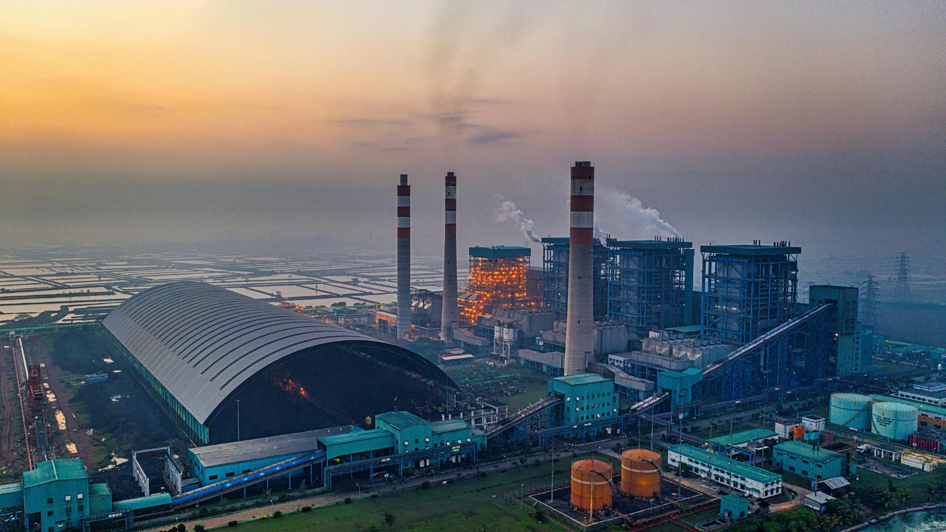 Aerial landscape view of a massive industrial power generation plant and arched storage facility illuminated against a colorful sunset sky.