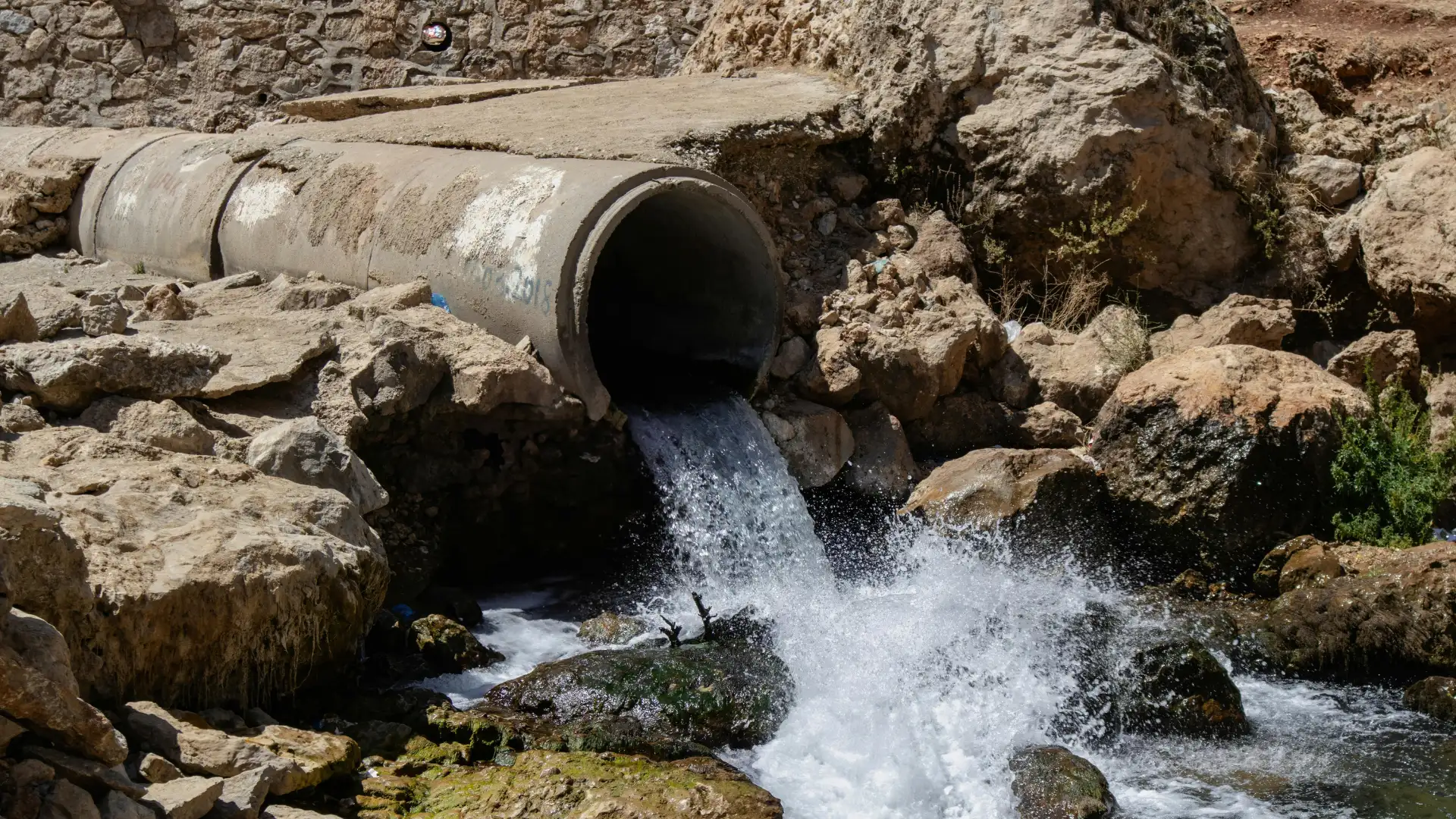 Concrete drainage pipe discharging treated wastewater effluent into a natural rocky stream environment.