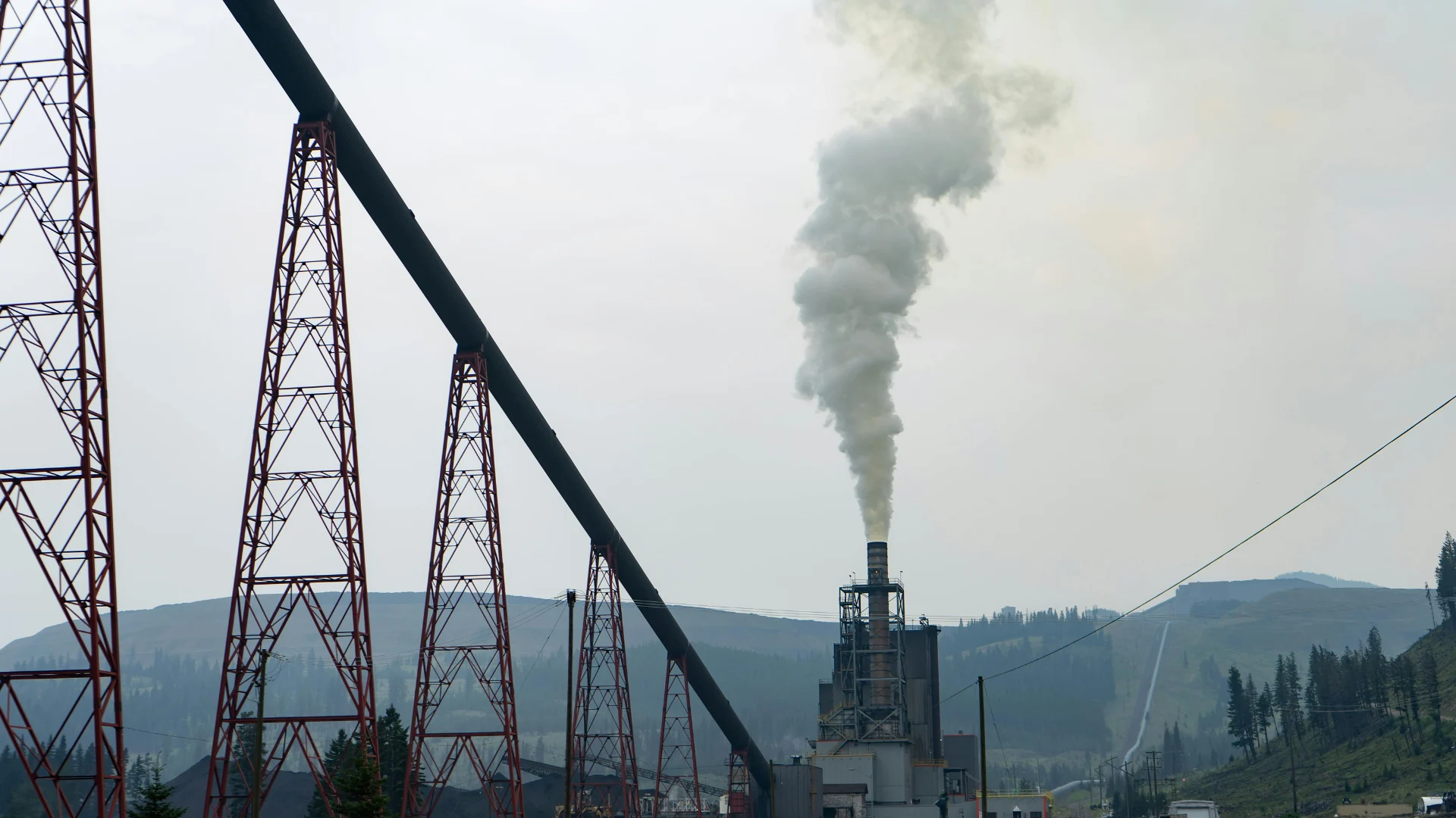 Large industrial mining and processing facility featuring a long elevated conveyor belt system and a smokestack releasing emissions into the sky.