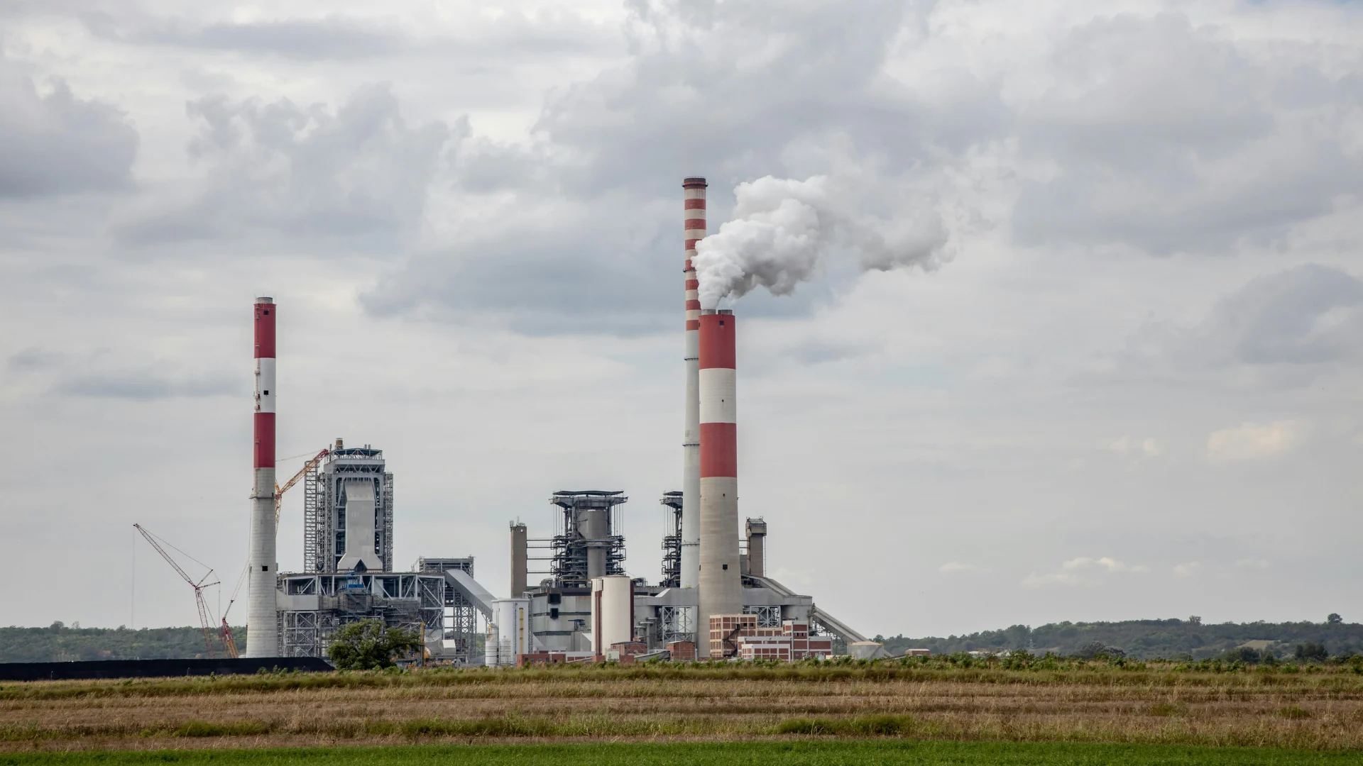Tall red and white smokestacks emitting steam and smoke at a large industrial power generation plant in a rural landscape.