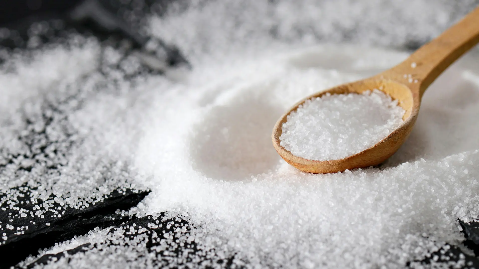 Close-up of a fine white crystalline powder or chemical granules resting on a wooden measuring spoon against a dark, textured slate background.
