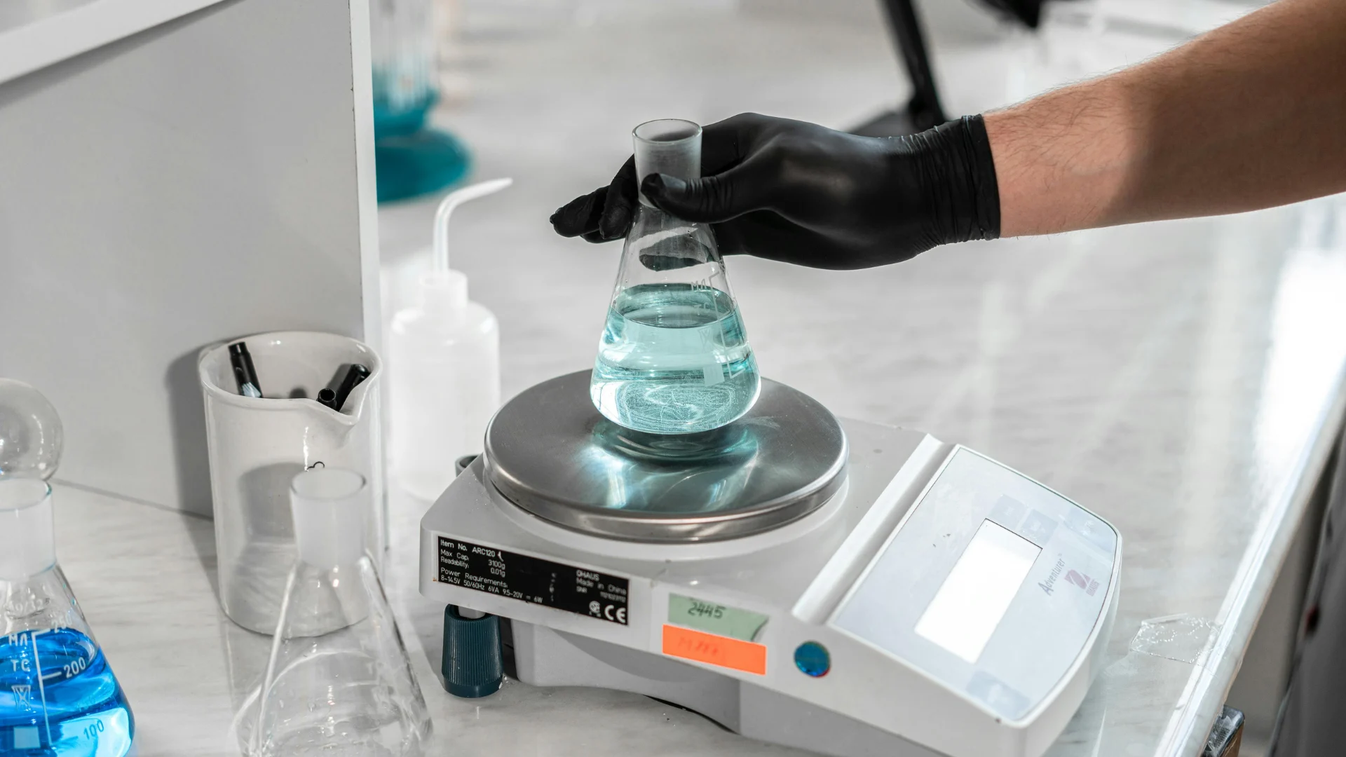 Technician weighing a glass Erlenmeyer flask of blue chemical liquid on a digital scale during laboratory quality control testing.