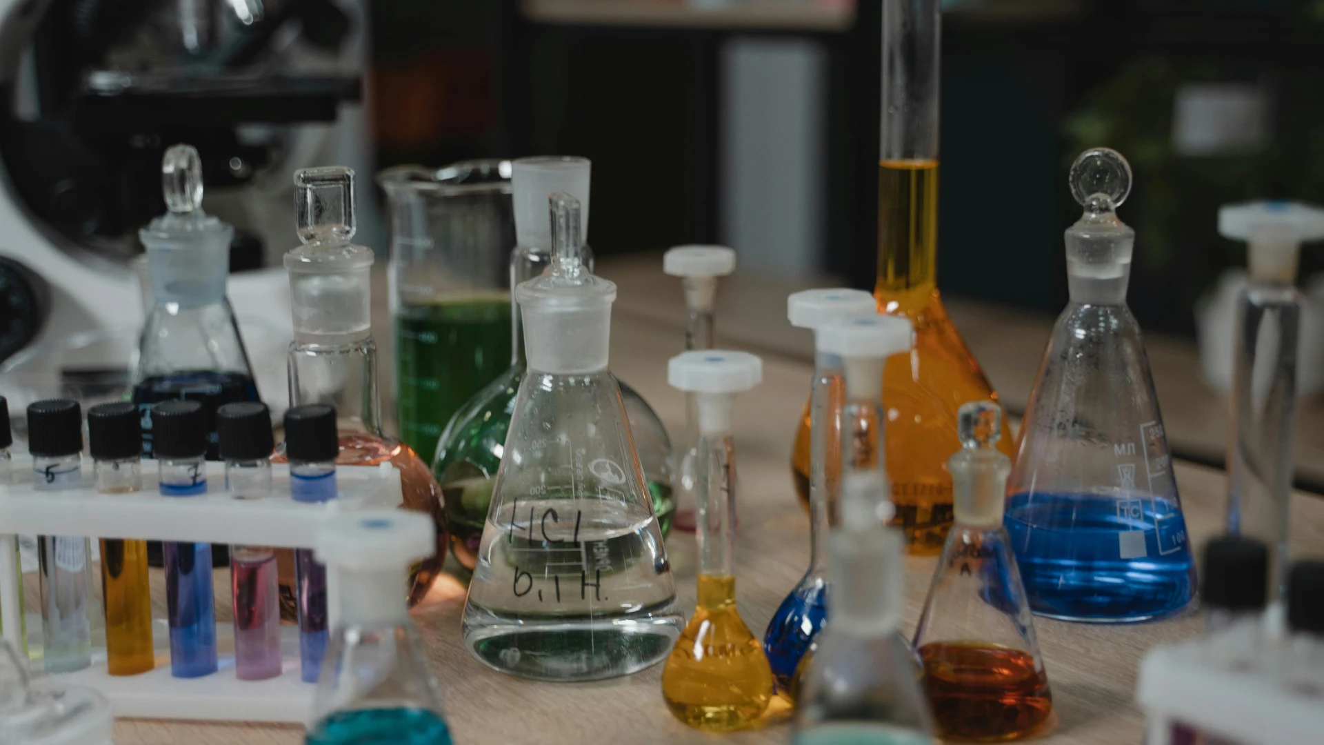 A chemistry laboratory workspace featuring multiple glass Erlenmeyer flasks, beakers, and test tubes filled with brightly colored liquid chemical solutions.