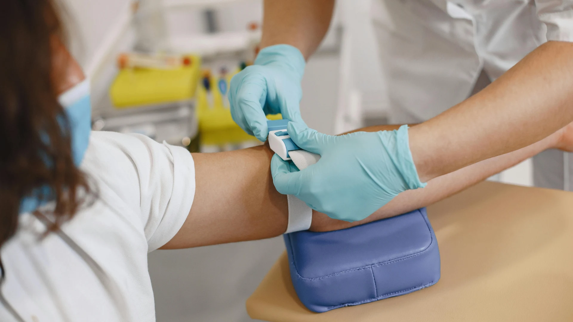 A healthcare professional wearing protective blue gloves applies a tourniquet to a patient's arm in preparation for a clinical blood draw and laboratory testing.