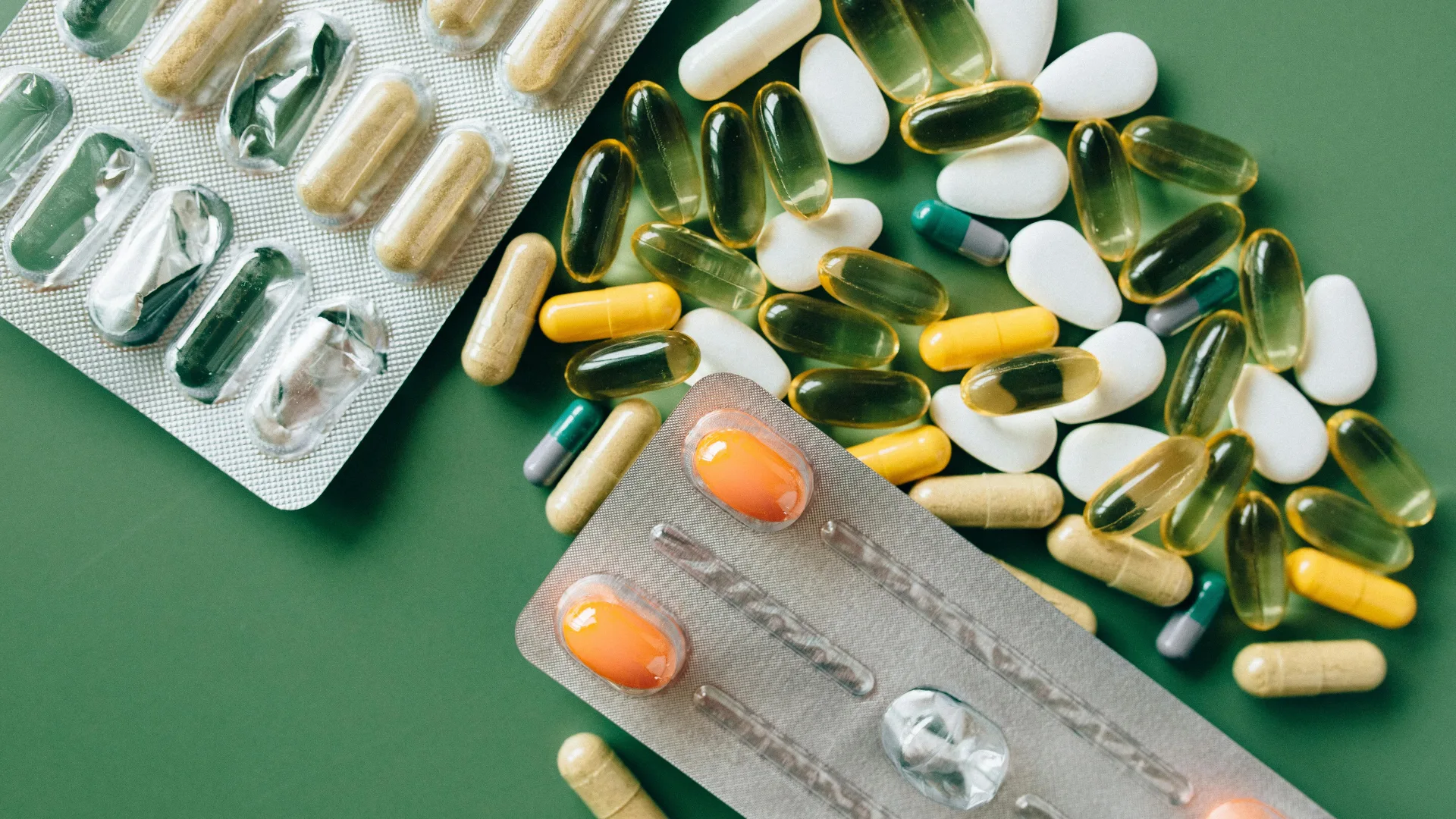 Assorted pharmaceutical medicine, including yellow and green liquid gel capsules and white tablets, scattered next to foil blister packs on a solid green surface.
