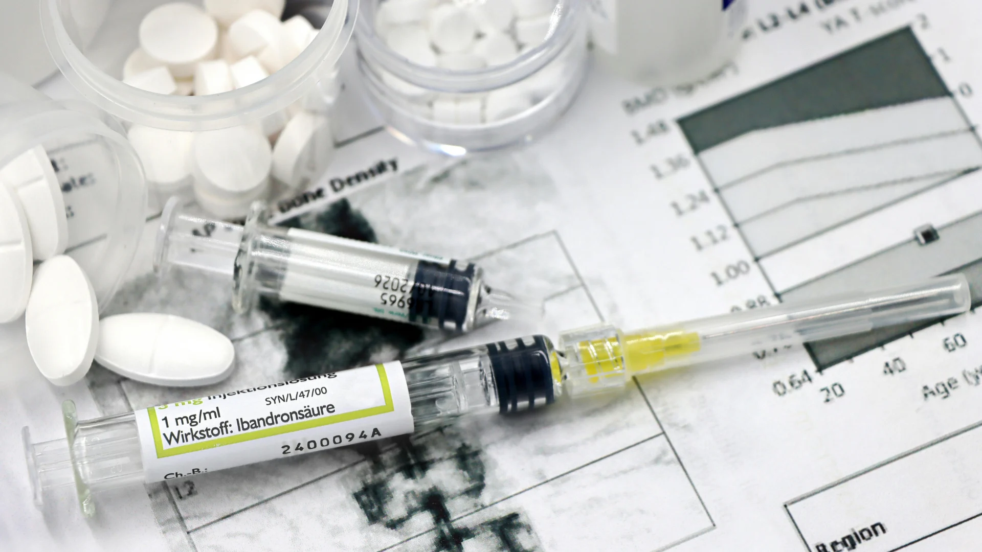 Medical syringes, white prescription pills, and medication vials resting on top of printed clinical patient charts and medical graphs representing pharmaceutical research.