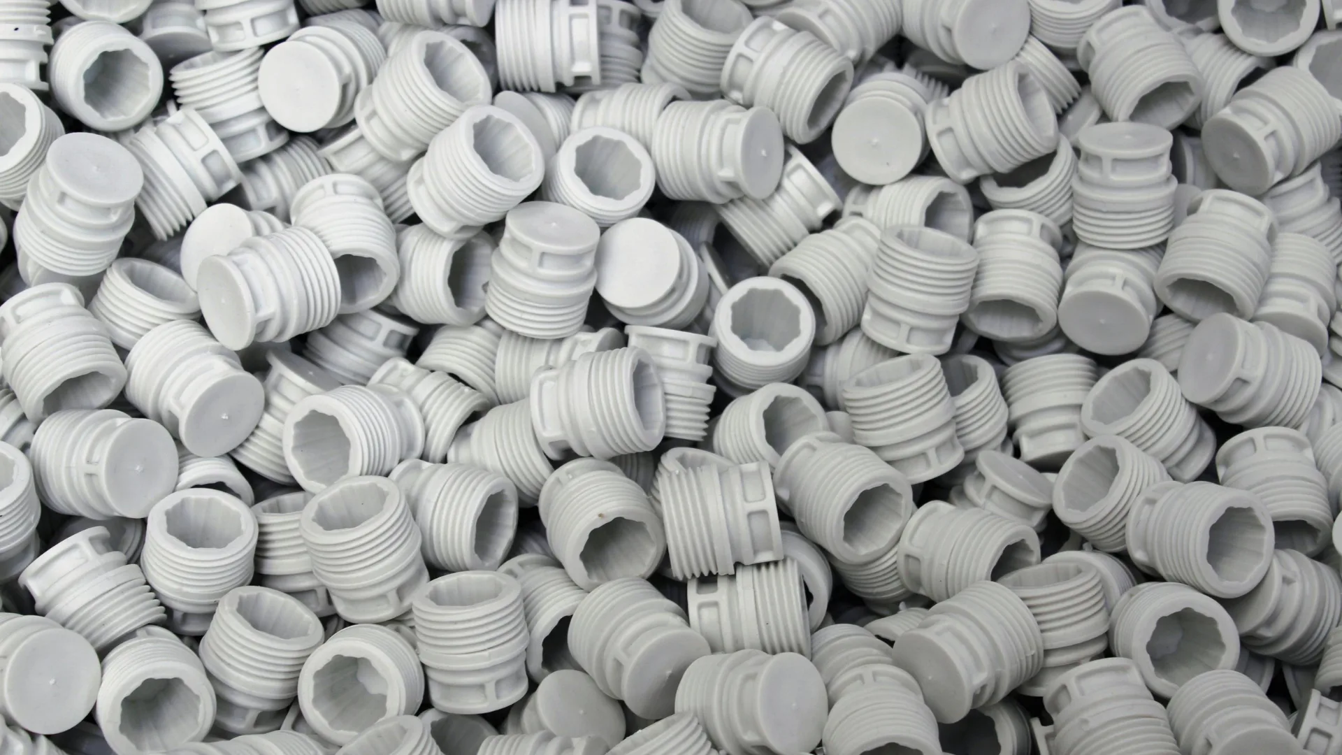 A large pile of manufactured grey plastic threaded caps and plugs, showcasing high-volume industrial plastic injection molding and polymer compounding.
