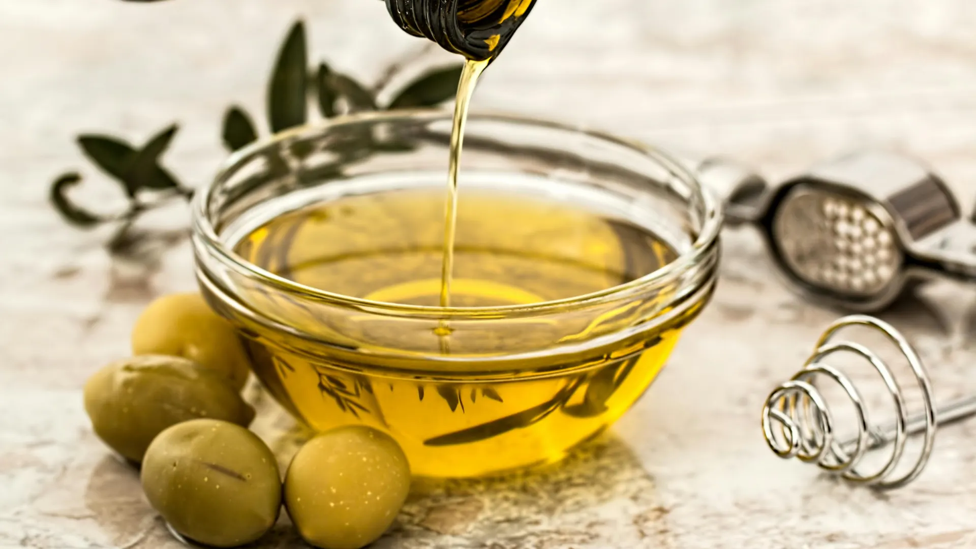 Golden extra virgin olive oil being poured from a dark glass bottle into a clear bowl, accompanied by fresh green olives and a metallic garlic press on a marble countertop.