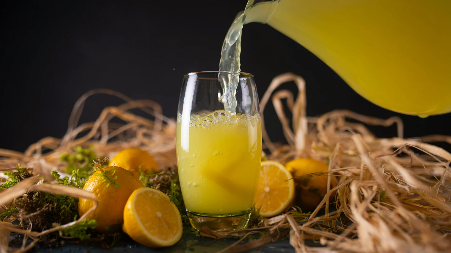 Fresh, vibrant yellow lemon juice being poured from a pitcher into a glass, surrounded by whole lemons, rustic straw, and natural green moss.
