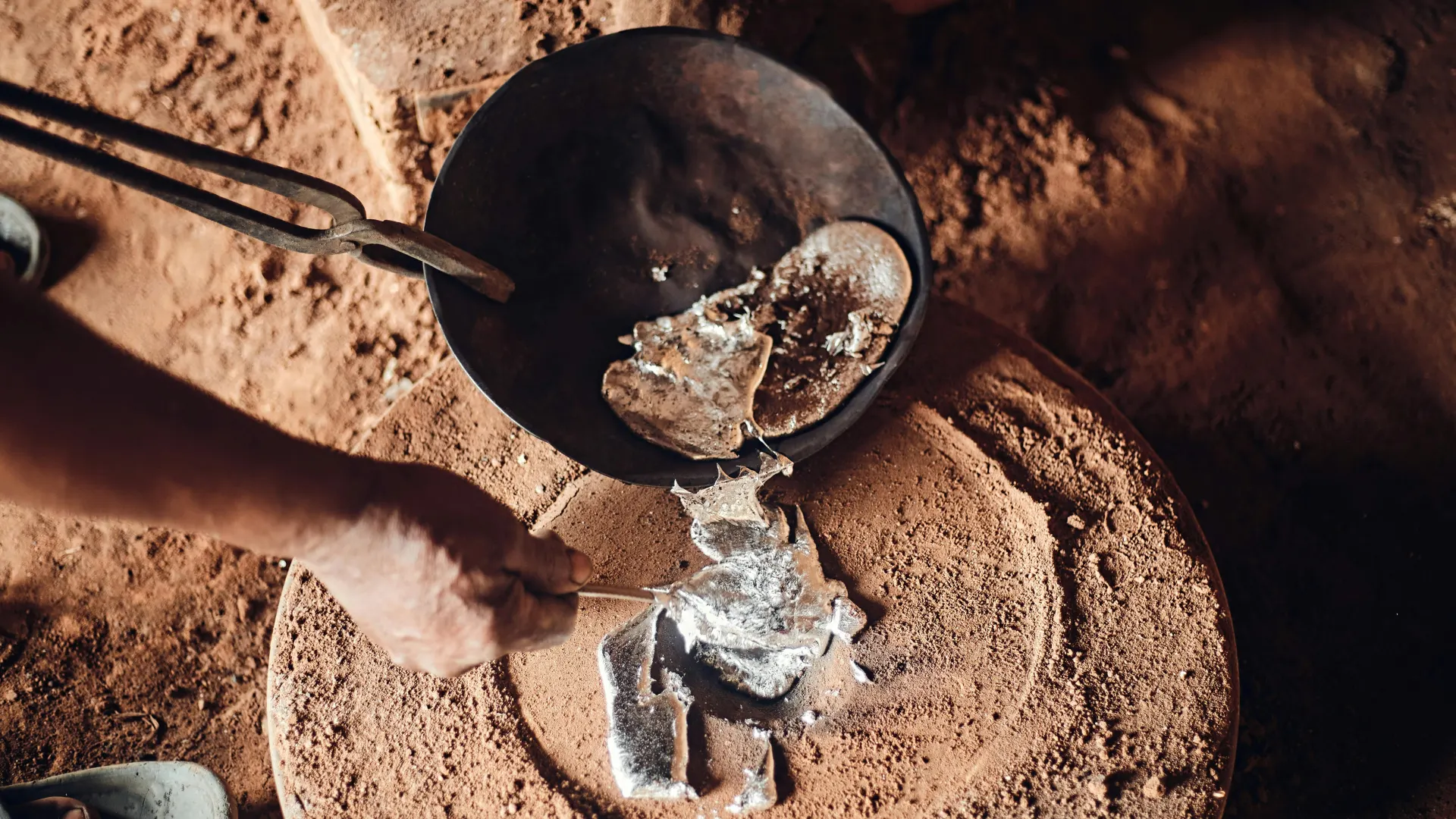 A foundry worker using metal tongs to carefully pour glowing, liquid silver-colored metal from a dark crucible onto a textured sand casting bed.