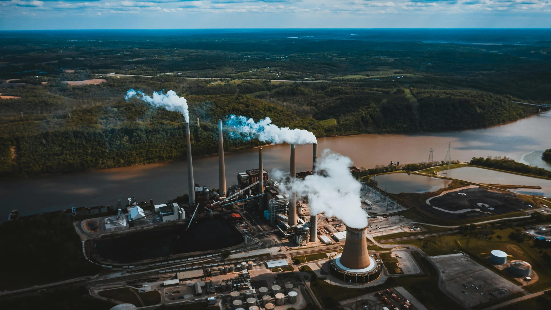Aerial view of a large-scale industrial power plant situated along a brown river, with tall smokestacks releasing thick white plumes of steam into the air.