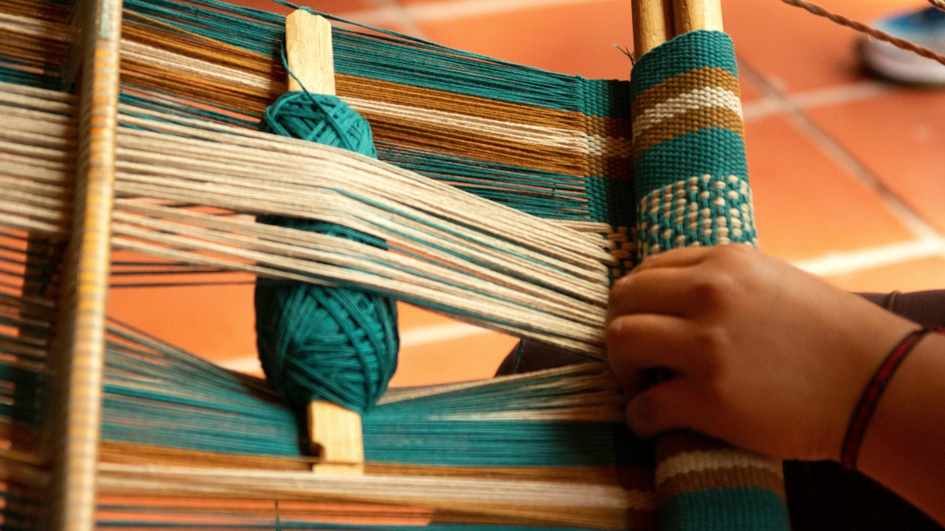 Close-up of a person weaving bright green and brown synthetic threads on a wooden handloom, representing textile production and commercial fabric dyeing.